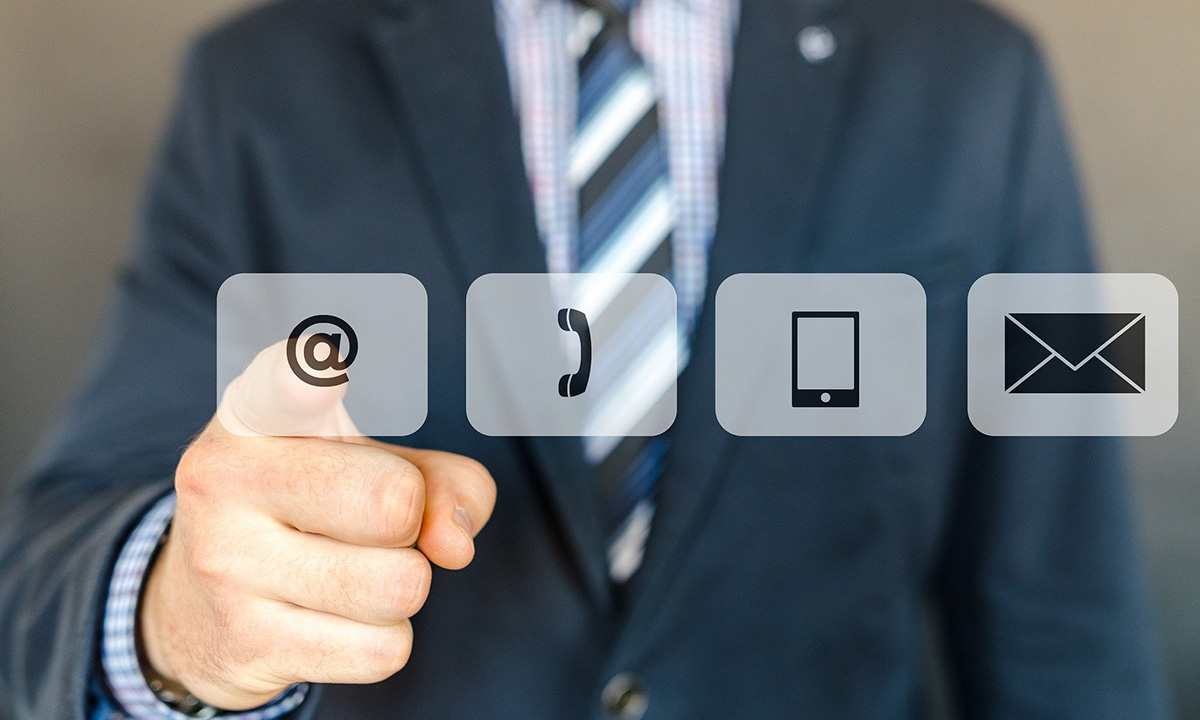 Das Bild trägt den Titel "Contact us". Es zeigt einen Mann im Anzug, der hinter einer Glasscheibe steht und auf ein darauf befindliches @-Symbol zeigt. Rechts daneben sind noch die Symbole eines Telefonhörer, eines Smartphone und eines Briefumschlag auf der Glasscheibe abgebildet.